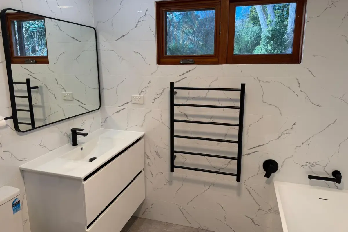 The same Katoomba bathroom after renovation, tiled floor to ceiling in marble-look tiles with a wall-hung vanity, a black heated towel rail and a black mixer over the new bath