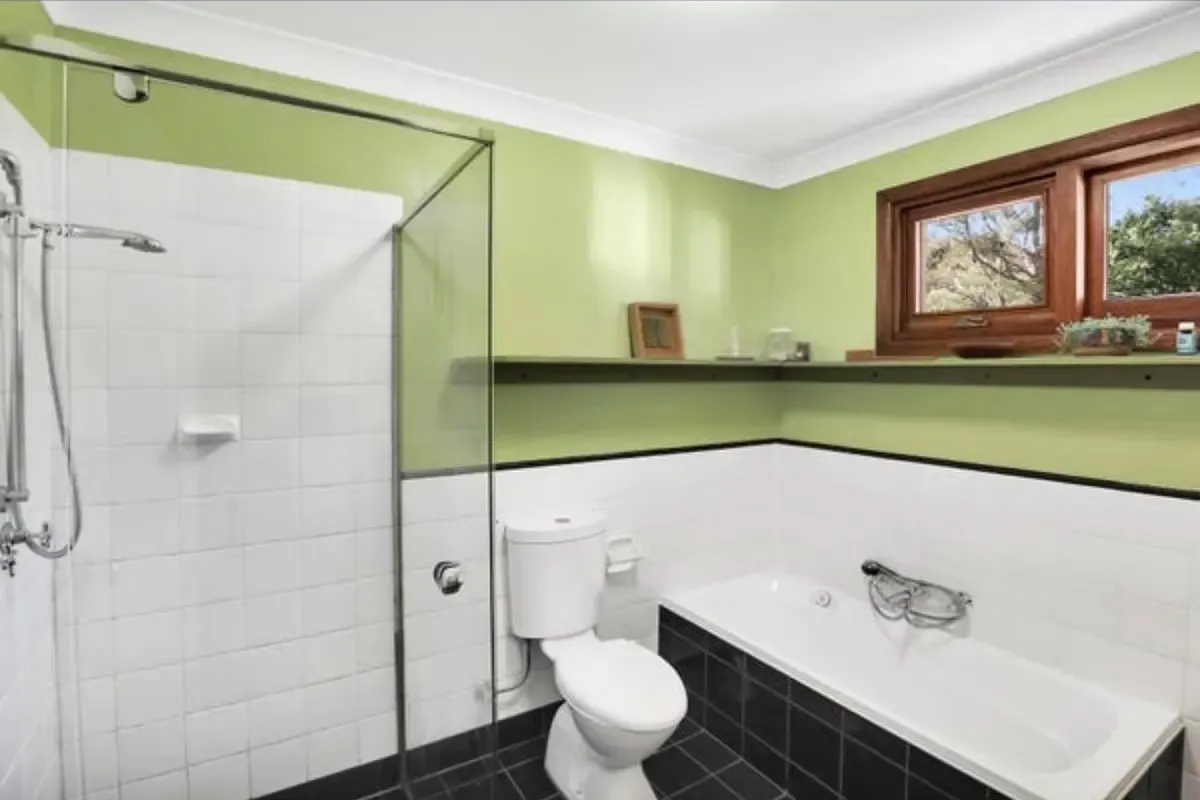 The Katoomba bathroom before Quick Mick’s Plumbing renovated it, with green walls, white square wall tiles, a dark tiled floor and an old drop-in bath under the timber window